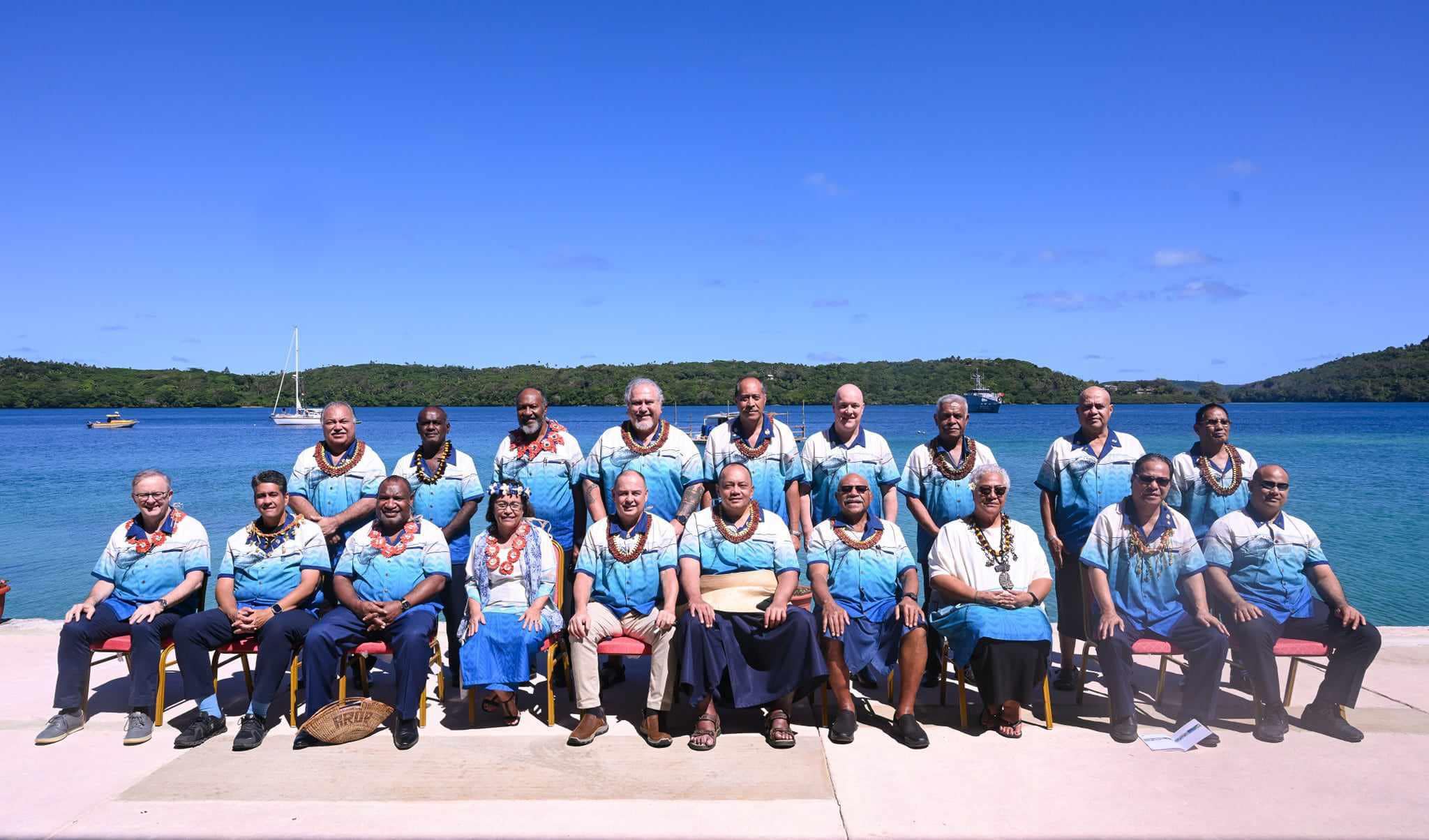 A large group of men and women seated in matching shirts and leis, Sail boats are visible in the bay in the background.