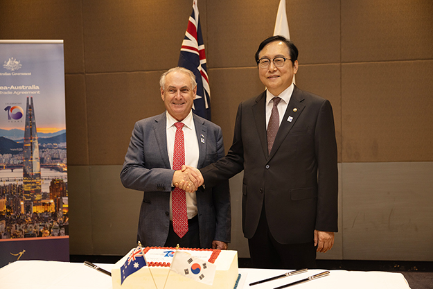 The Hon Don Farrell, Minister for Trade and Tourism, Australia and Dr Cheong Inkyo, Minister for Trade, the Republic of Korea, are smiling and shaking hands while standing behind a cake that is celebrating the 10-year anniversary of the Korea-Australia Free Trade Agreement. An Australian and a Korean flag are behind the ministers.