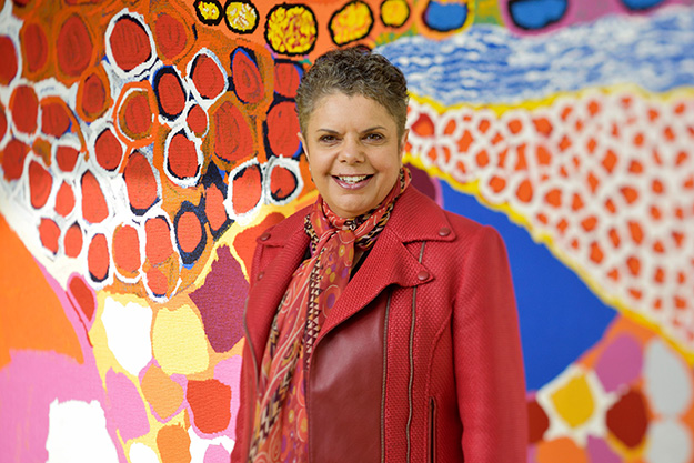 A First Nations woman wearing a red jacket, and a red scarf stands in front of a vibrant First Nations tapestry.