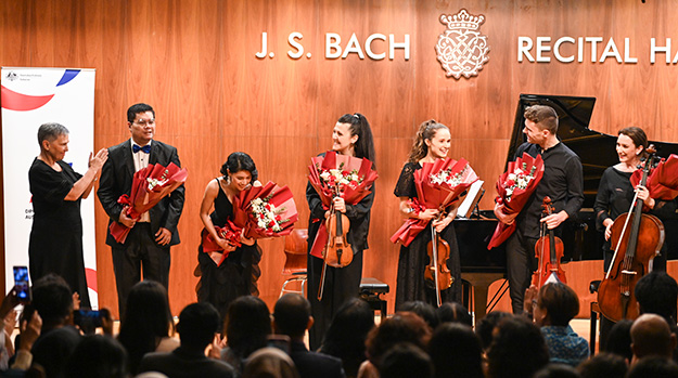 A small group of musicians take bows after their performance while holding their instruments and bunches of flowers.