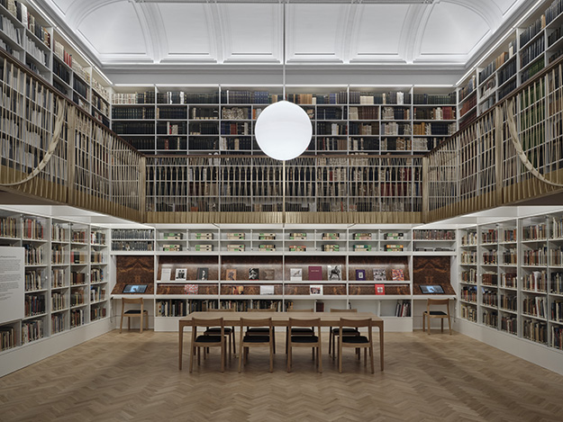 A large, open plan library with books lining the wall and a large, single pendant light hangs from the centre of an arched ceiling.