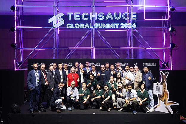 A large group of people gathered on stage, lit with purple-neon lighting. Sign in the background 'Techsauce Global Summit 2024'.