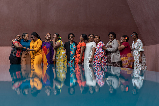 12 WDLP participants dressed in bright colours, closely lined up with their arms around each other and their reflection visible on the blue platform in front of them.