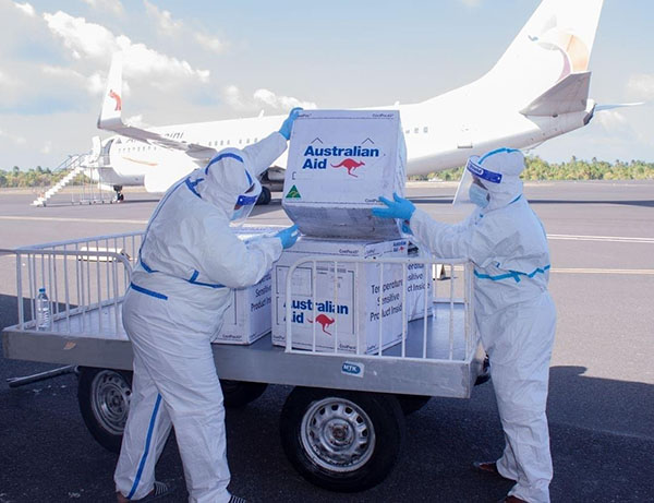 Two people in full PE gear unloading boxes of COVID-19 vaccines from a trailer. A plane is parked on the runway behind.