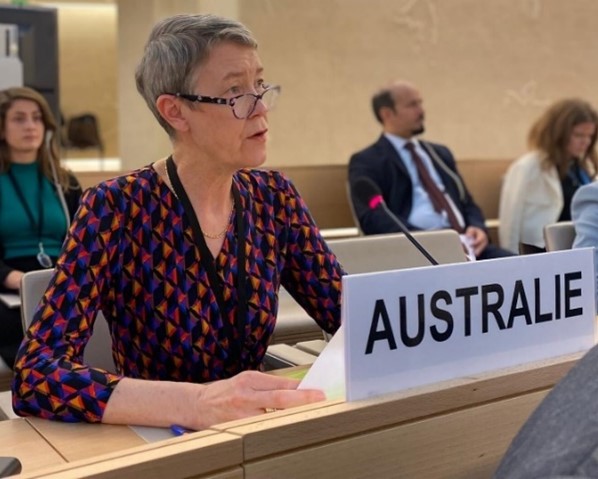 A woman sitting at a desk and speaking into a microphone. A name card with the word "Australie" is on the desk in front of her.