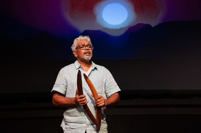 A man holding two boomerangs stands singing with an image of a full moon in the background.