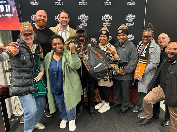 A group of people with NRL Wests Tigers merchandise, standing in front of a panel patterned with the team’s logo.