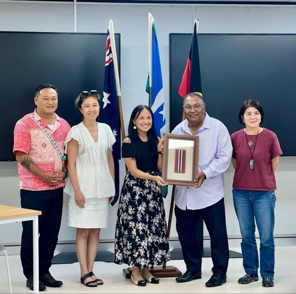 five people standing in front of Australian, Torres Strait Islands and Indigenous Australian flags, two people holding a framed picture.