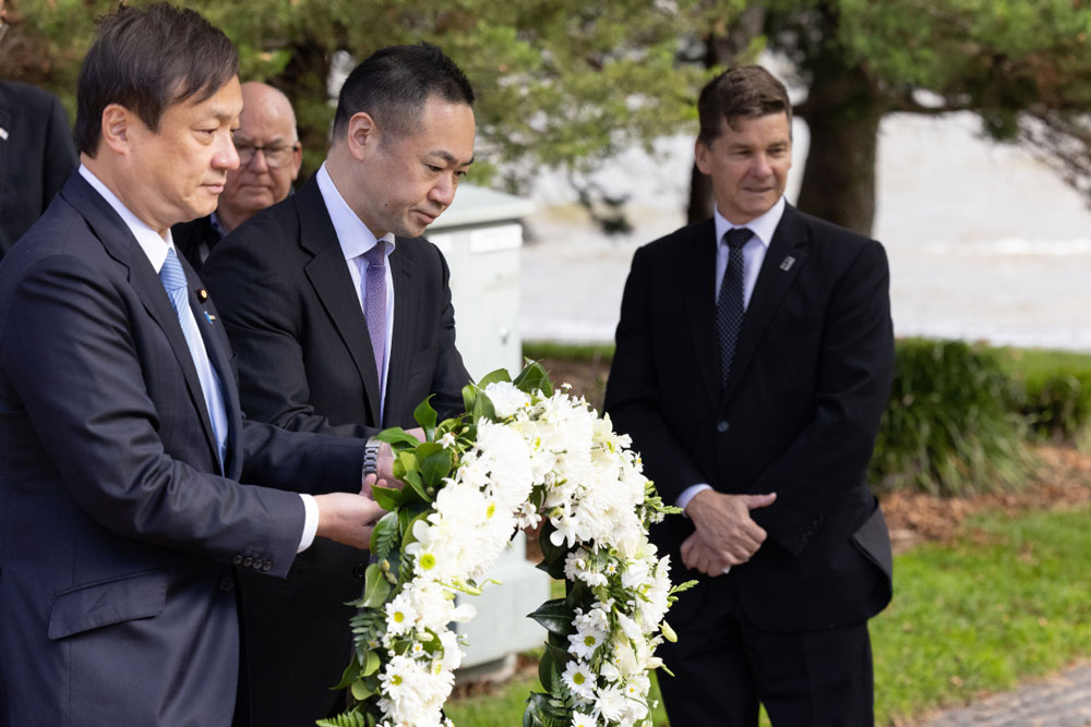 Two Japanese men laying a white floral wreath, watched by host Mr Smythe. In the background are trees and a lake.