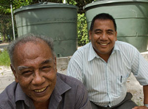 Two men from Kiribati are squatting down smiling. There are two large green water tanks in the background.