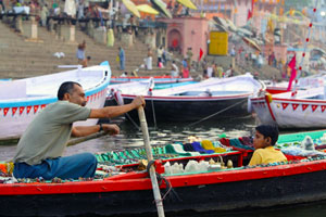 Man and younger boy are sitting in a boat on a river in front of a number of stationary boats. The man is holding the oars rowing. The boat is filled with colourful cloths. In the background on the bank of the river are a number of large steps with lots of people standing around.