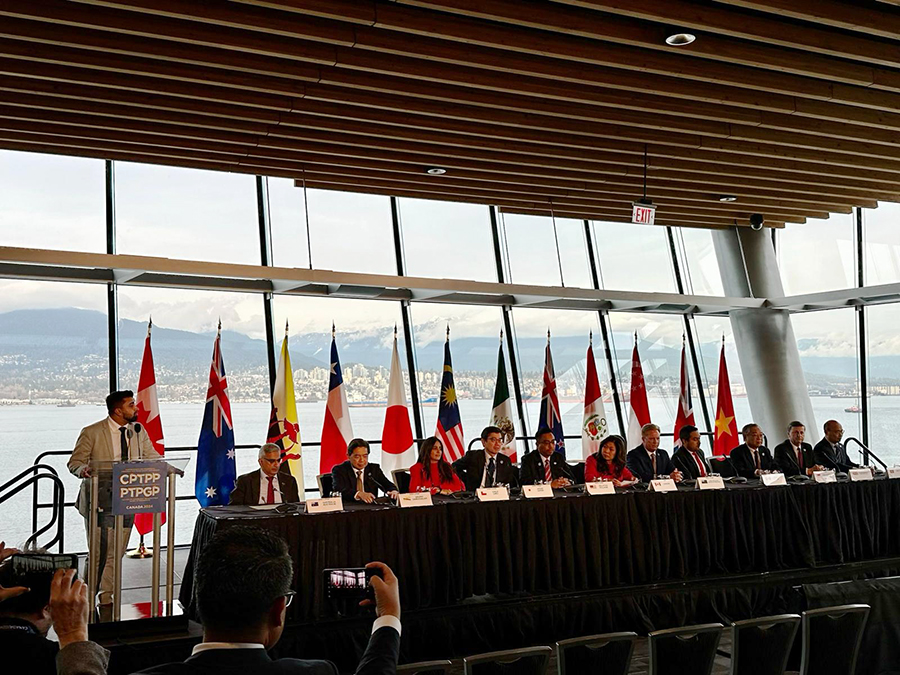 11 people seated behind a long table, with country flags in the background. A man MCs from a lectern to one side. View of a lake and mountains through large windows.