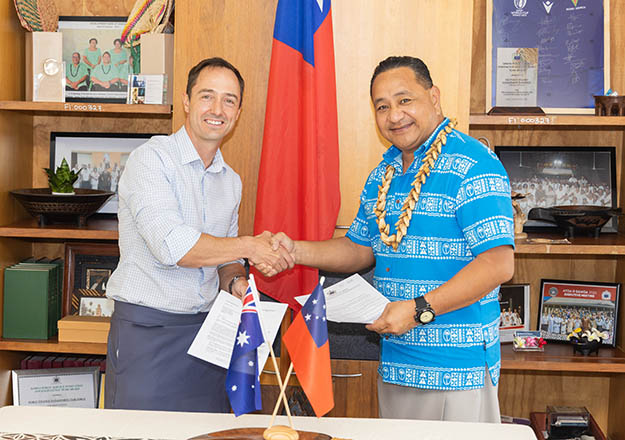 Two men shaking hands with Australian and Samoan table flags in front of them and a large Samoan flag behind them.