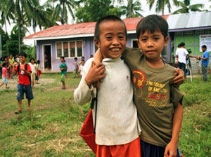 Two young Filipino boys with arms around each other smiling in foreground at school. A number of other children are playing in the background.