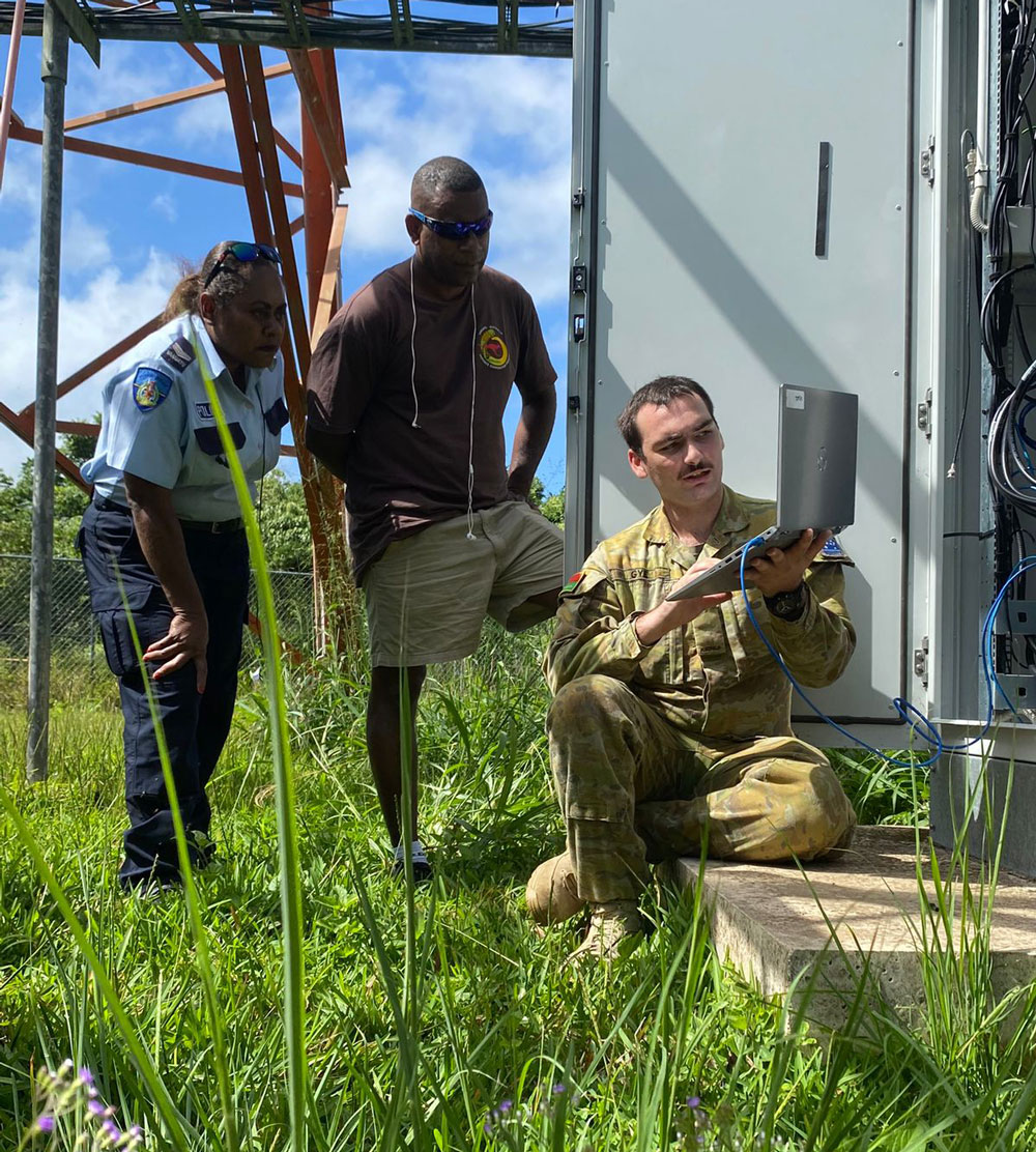 Photo of Australian Defence officer repairing a radio tower whilst two Vanuatu people watch and learn. 