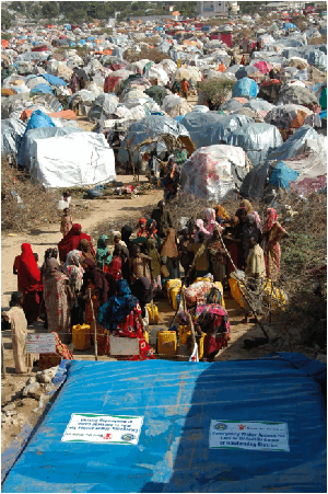 Two dozen Kenyan women are filling large plastic bottles with water from a large blue bladder displaying the Save the Children logo. In the background a large number of makeshift tents stretch into the distance.