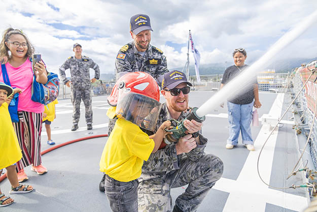 A young Samoan child in a helmet spraying water from a hose with the help of a uniformed Australian Defence personnel aboard a naval vessel, watched on by other Defence personnel and members of the public.