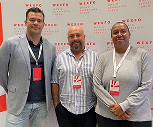 Three people standing together wearing lanyards, in front of a WEXFO media wall.