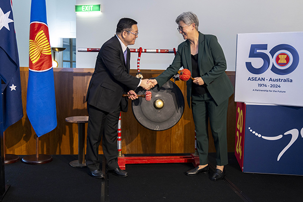 Two people shaking hands in front of a gong. @ flags are to their left and a sing to their right.