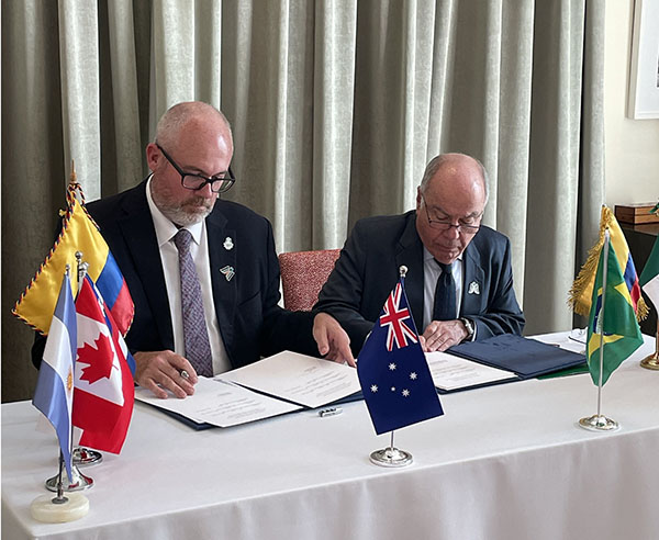 Two men sitting at a table signing documents surrounded by flags