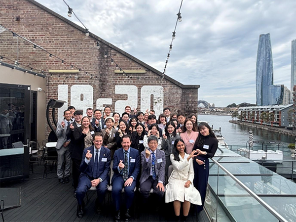 Group photo of formally dressed individuals on an outdoor deck overlooking a waterfront, with a brick building marked '19-20' and modern high-rises in the background—likely taken during a professional or formal event.