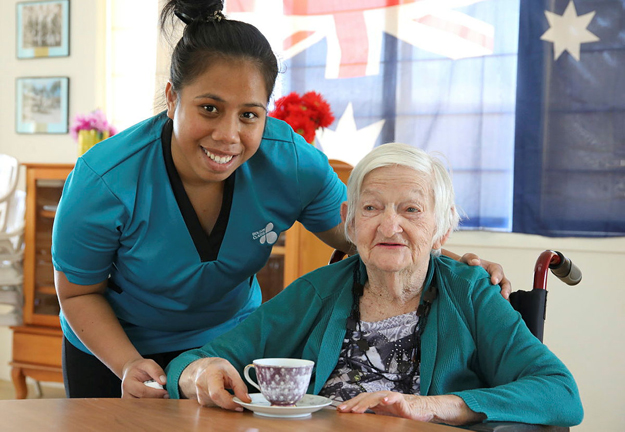 A nurse stands behind an aged care resident at a table in a wheelchair, with a cup of tea.