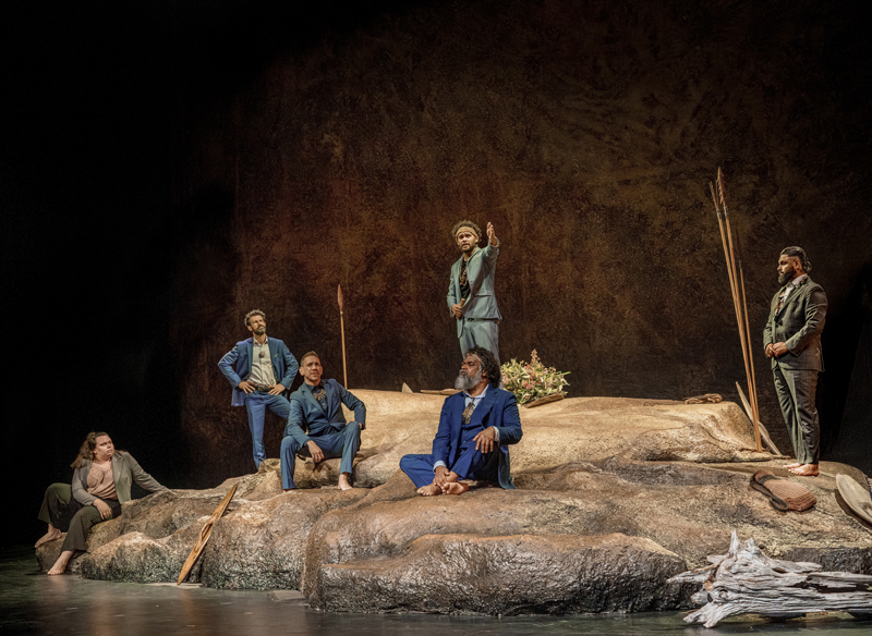 Six people posing on stage, barefoot, wearing suits on a set designed to look like rock. Indigenous spears and shields are placed around the set.