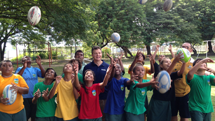 Parl Sec Matt Thistlethwaite throwing AFL footballs in the air with a crowd of smiling children.