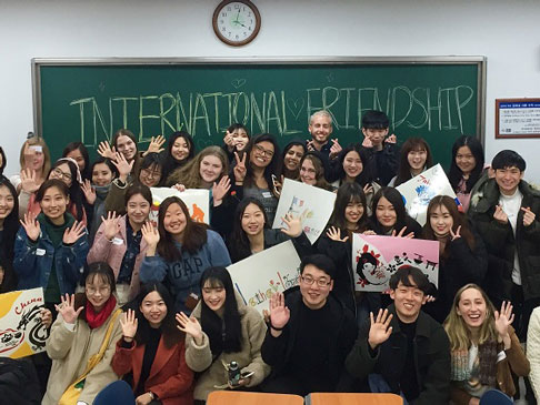 Class of students waving. The chalkboard at the back of the class has International Friendship written on it.