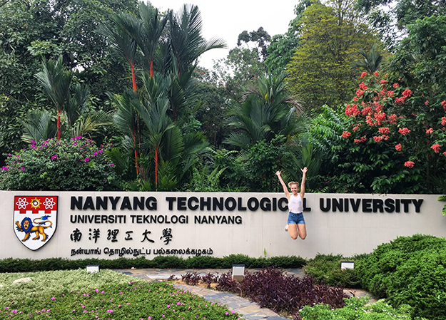 A woman wearing denim shorts and a white singlet jumping with arms raised in front of a Nanyang Technological University Singapore sign. Dense tropical vegetation in the background.