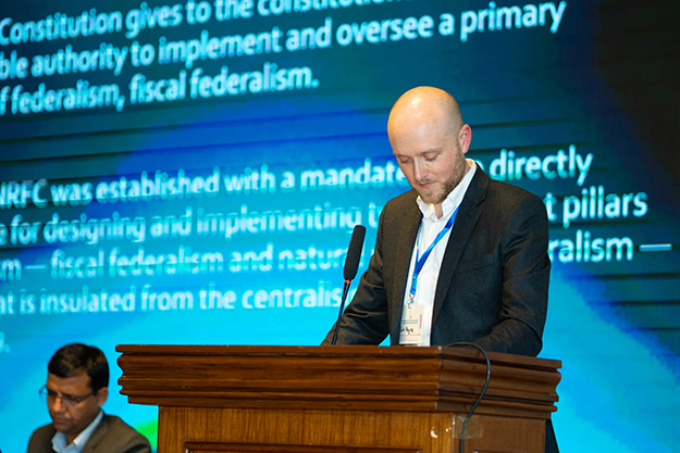 Man at a podium presenting at a conference with a PowerPoint slide showing in the background