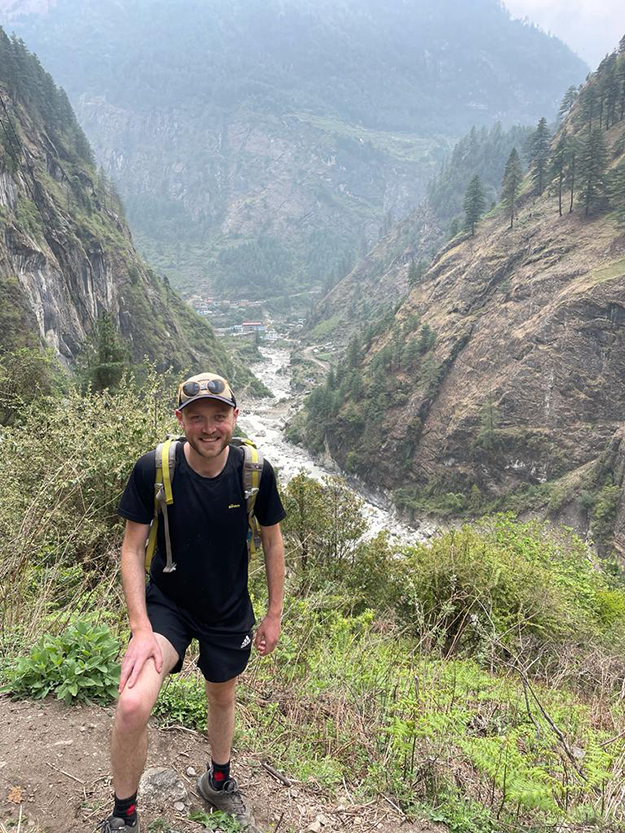 Man on a mountain side with a backpack, hat and glasses. River can be seen below with mountains off into the background. 