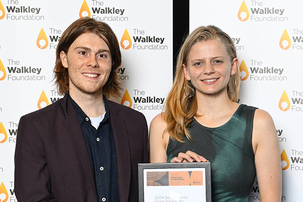 Man and a women standing in front of a Walkley Foundation media wall, holding a framed certificate.