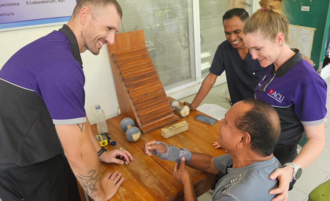 Three people standing around a man seated at a table with a brace on his right arm.