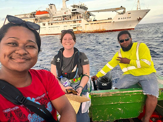 Three people in a small boat with a large boat behind in the distance