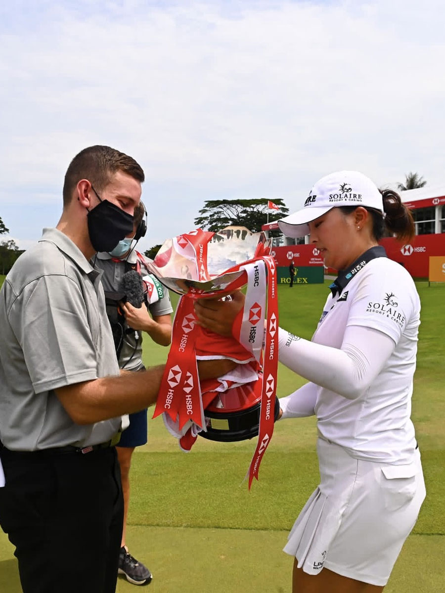 Photo of Jack Steed passing a trophy to Jin Young Ko on a golf course.