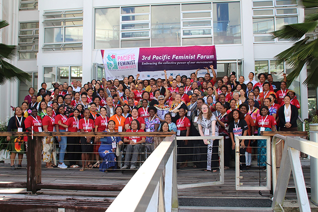 A large group of women posing for a group photo with a banner 'pacific feminist forum 2023: 3rd pacific feminist forum, embracing the collective power of movements'.