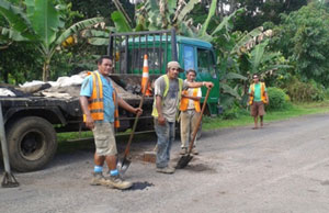 Three Samoan workmen standing together on a road carrying  spades in front of their work truck. Another workman in standing in the background.