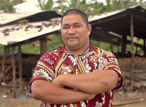 Samasoni standing on front of hut covered with rubble.