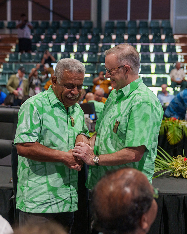 Alt text: two men in green shirts shake hands and smile.