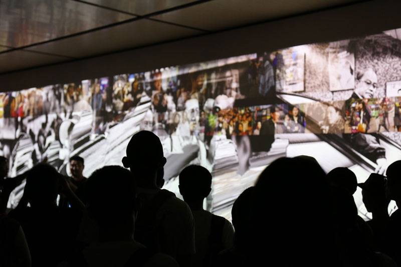 People in a dark room looking at a wall with a broadcast on it