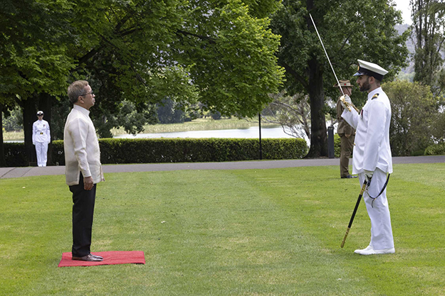 A man standing on a square of red carpet, facing a man in military dress uniform, holding up a sword.