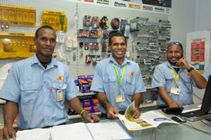 Three men in PNG standing behind store counter in blue uniforms. Man on the far right is talking on the phone. Behind the men is a display of store merchandise.