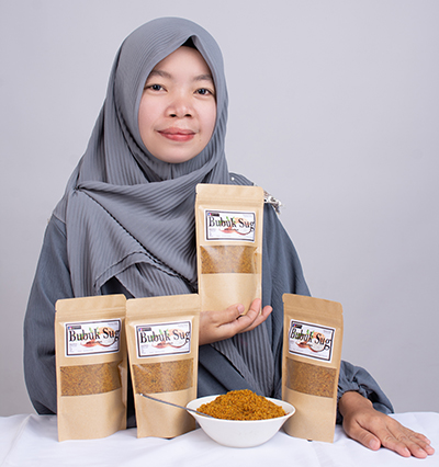 A woman wearing a hijab holds a packed of bobuk, with more packages on display