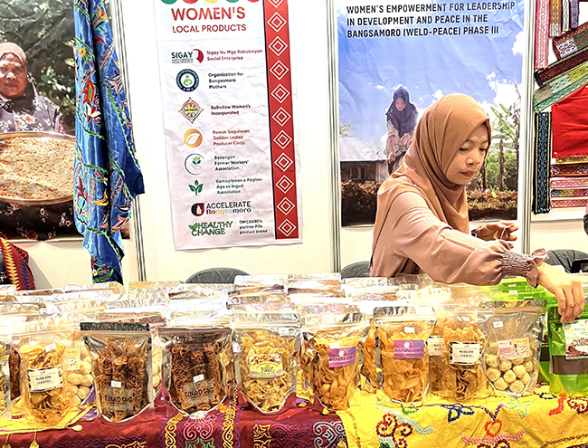 A woman wearing a hijab behind a market stall displaying colourful spice and food jars.