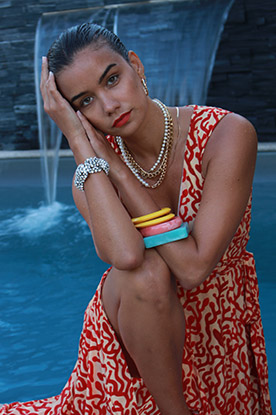 A model poses in front of a pool and waterfall, wearing bright coloured bangles, beaded necklaces and a read-patterned dress. 