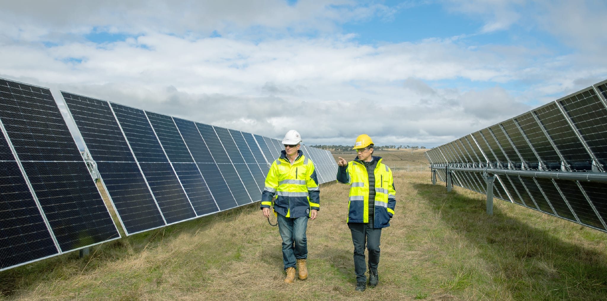 Two men in helmets and high-visibility jackets walking between two solar panels.