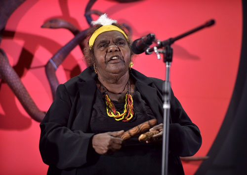 Senior Elder Ms Inawinytji Williamson sings Seven Sisters on a stage with clap sticks.