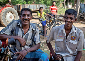 Two Sri Lankan men are sitting in the foreground smiling. The man on the left is resting on a bike. There are two boys in the back ground on bikes. There is a red tractor to the left behind the two boys.