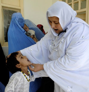 Female dentist holding a young boy's mouth open to inspect. Mother holding her sleeping baby waits in background.